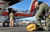 A smokejumper kneels beside an aircraft to do final checks before boarding. Two people stand near the aircraft doorway talking.
